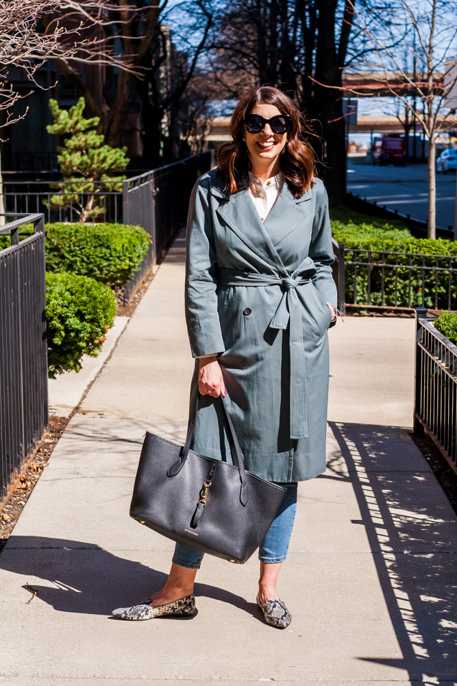 stylish woman smiling and wearing a unique trench coat, jeans, and holding a black bag